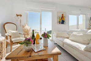A modern living room with a wooden table, white sofa, and a large window offering a city view.