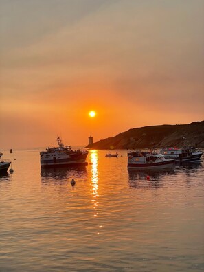 Un port au coucher du soleil, avec des bateaux amarrés, une eau calme et un phare à l’horizon.