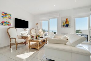 Ein modernes Wohnzimmer mit einem weißen Sofa, einem Holztisch, einem Korbstuhl und einem an der Wand befestigten Flachbildfernseher.