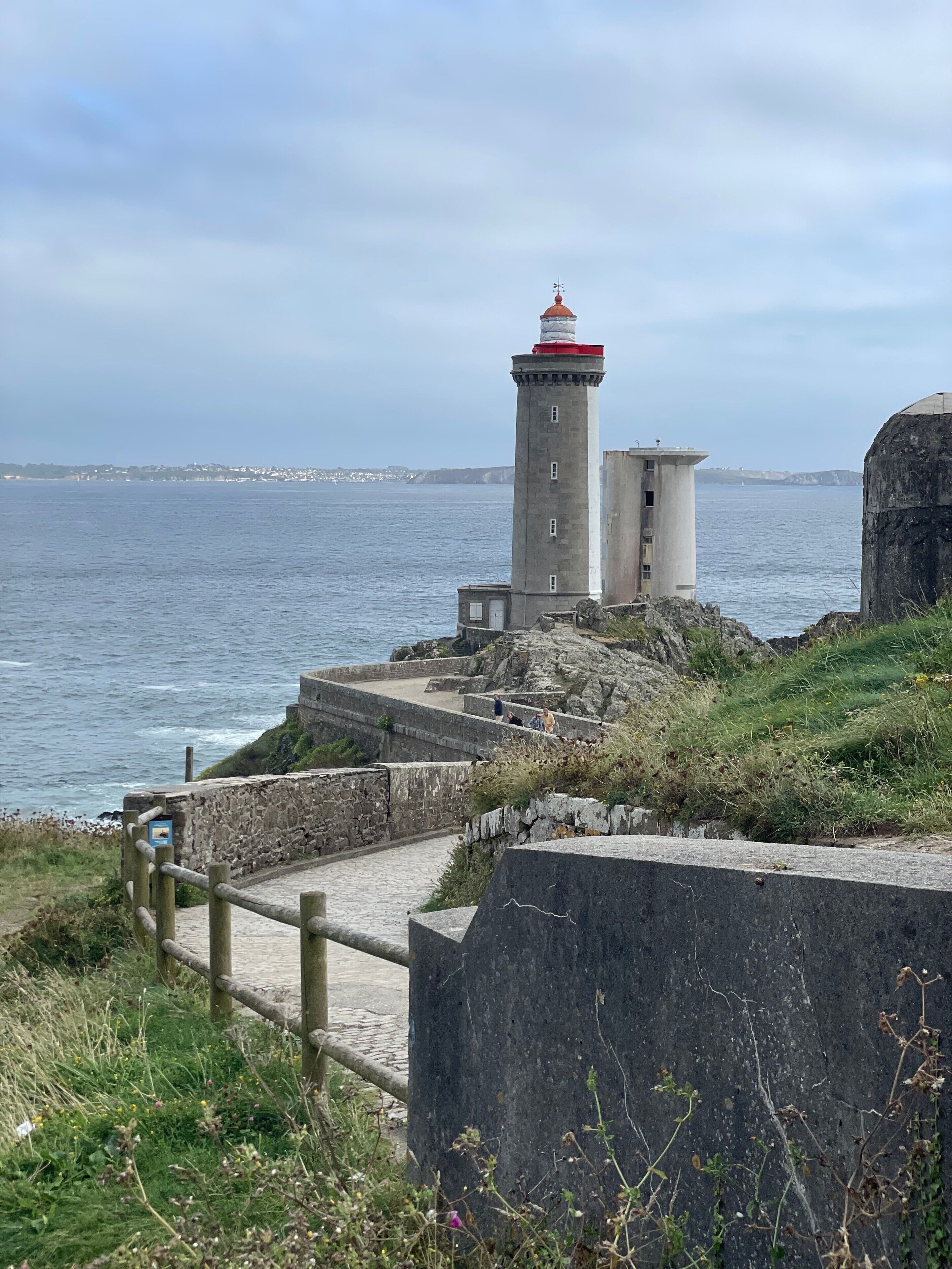 A lighthouse with a red top, situated on a rocky coastline with a paved walkway leading to it.