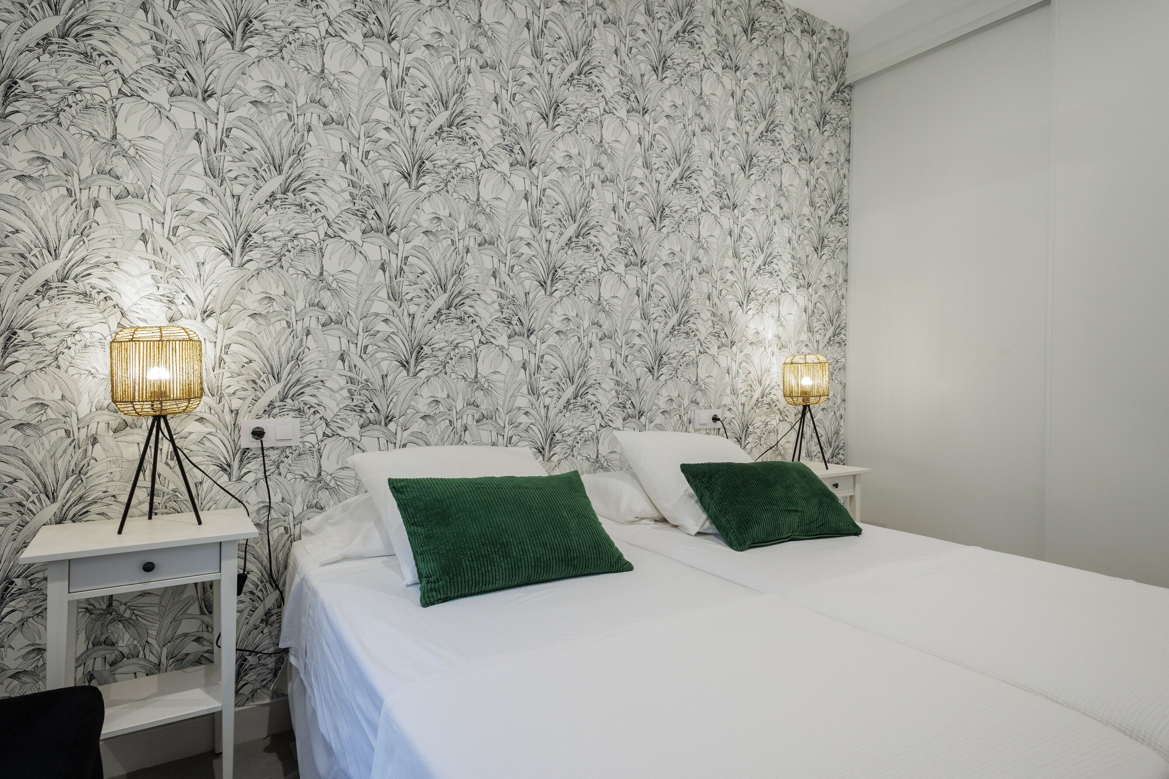 Un dormitorio con papel tapiz floral, dos mesitas de noche con lámparas, una cama con ropa de cama blanca y almohadas verdes, y una mesita de noche blanca.