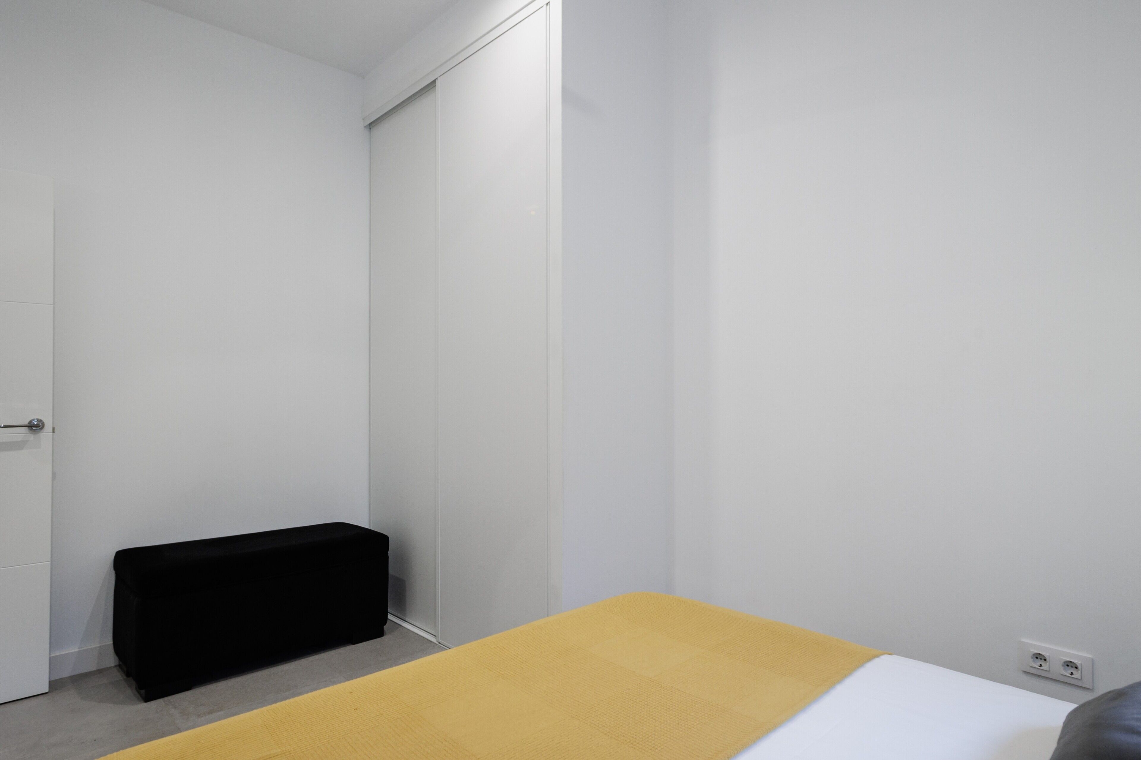 Una habitación con cama, banca, armario y toma de corriente.