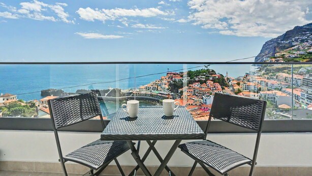 A balcony with a table and chairs, overlooking a coastal town with buildings and a clear sky. - Holiday Home 'Lobo's House' with Sea View, Private Terrace and Wi-Fi (Câmara de Lobos)