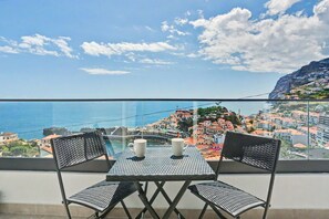 Un balcon avec une table et des chaises, donnant sur une ville côtière avec ses bâtiments et un ciel dégagé.