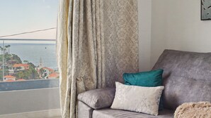 A living room with a sofa, a glass table, and a view of a coastal town. - Holiday Home 'Lobo's House' with Sea View, Private Terrace and Wi-Fi (Câmara de Lobos)