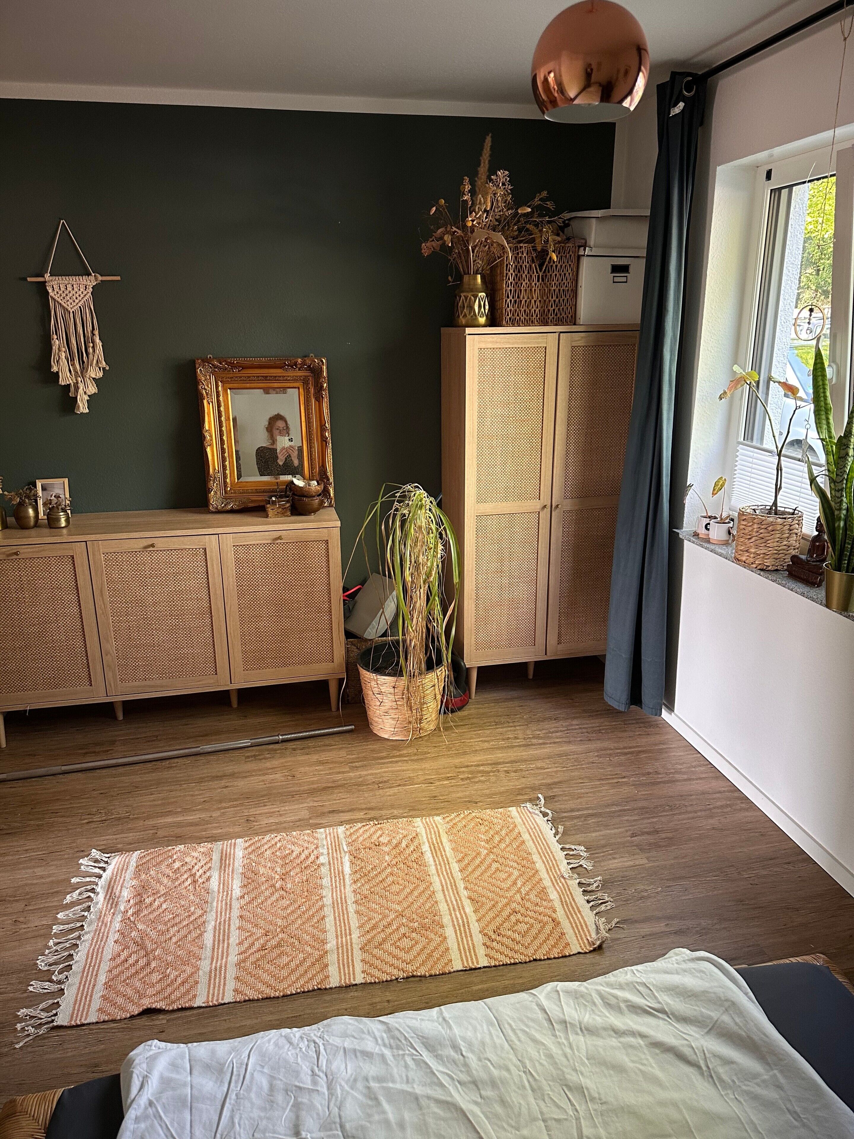 Ein Zimmer mit einem Holzkasten, einem Wandbehang aus Makramee, einem gerahmten Bild, einer Topfpflanze und einem Fenster mit Blick.