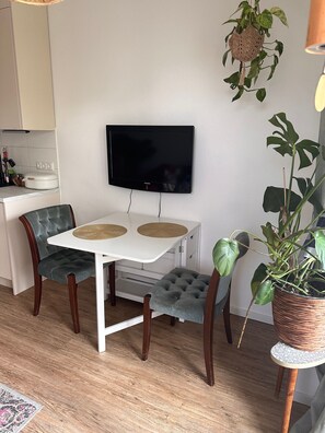 A dining area with a white table, two cushioned chairs, a television mounted on the wall, and a potted plant.