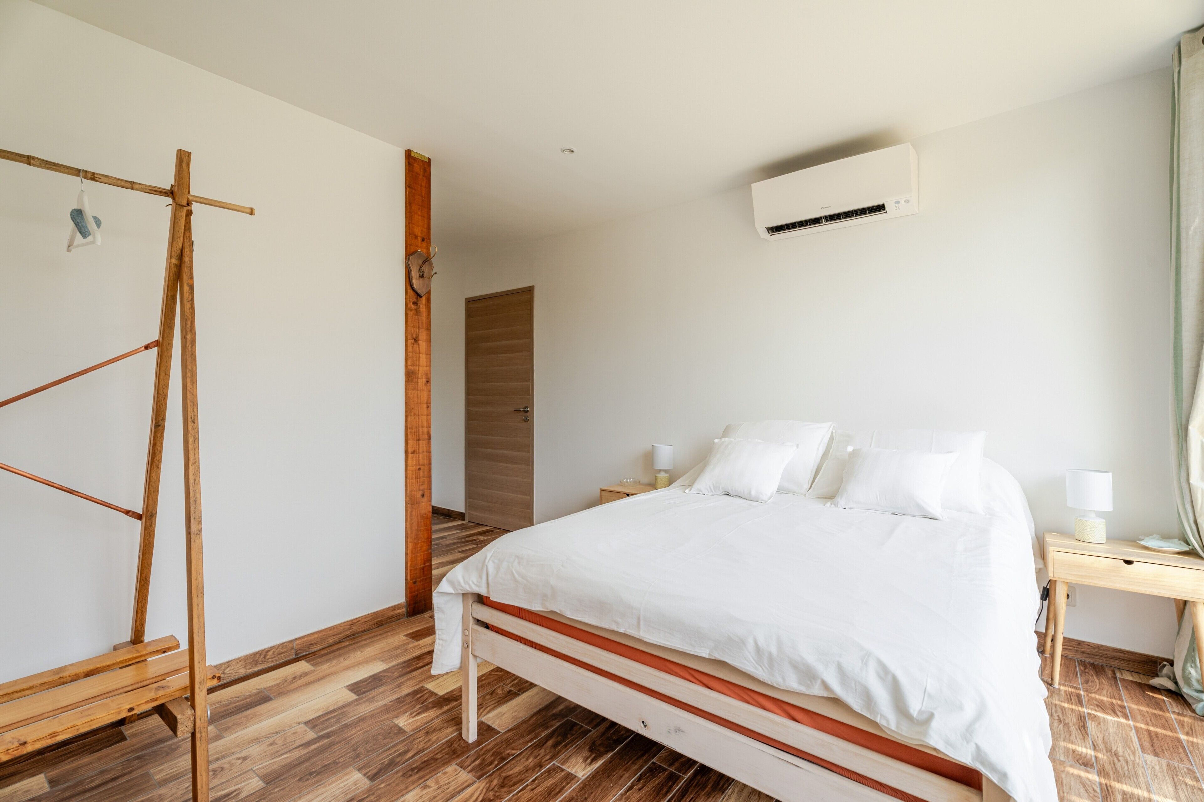 Una camera da letto con un letto, pavimento in legno, pareti bianche, un condizionatore d'aria, un armadio in legno e un comodino con una lampada.