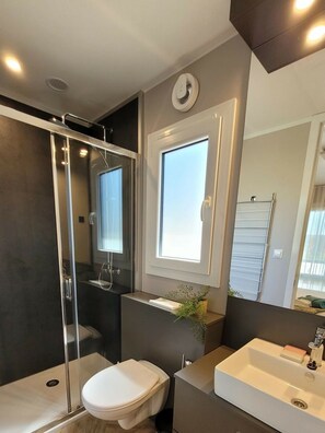 Een moderne badkamer met een douchecabine van glas, een toilet, een wastafel en een raam boven de spiegel.