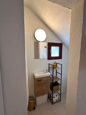 Casa de banho compacta com lavatório branco, espelho, pequena janela e um prateleira com artigos de higiene pessoal.