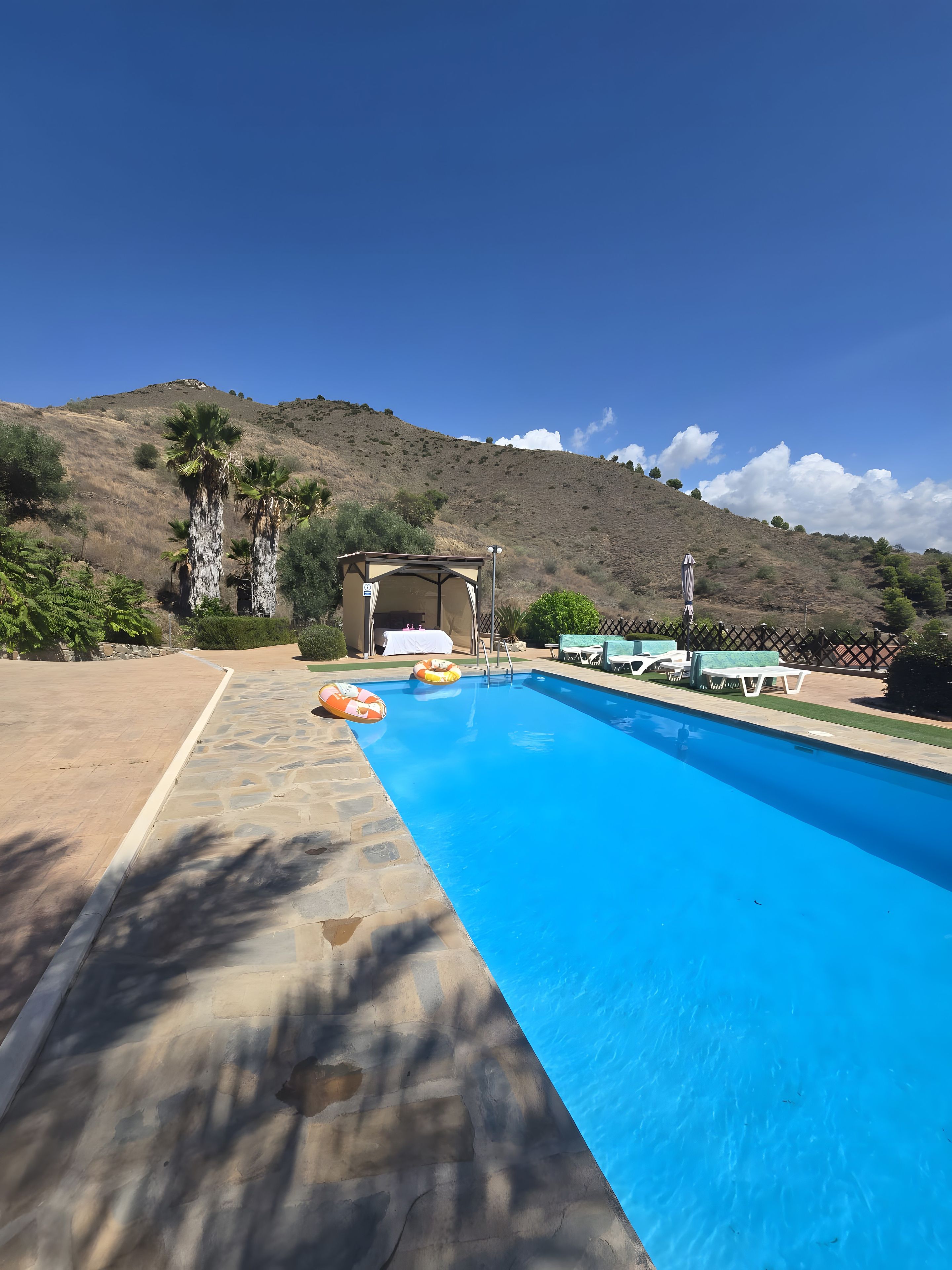 A swimming pool with lounge chairs and a covered area, surrounded by a hilly landscape.