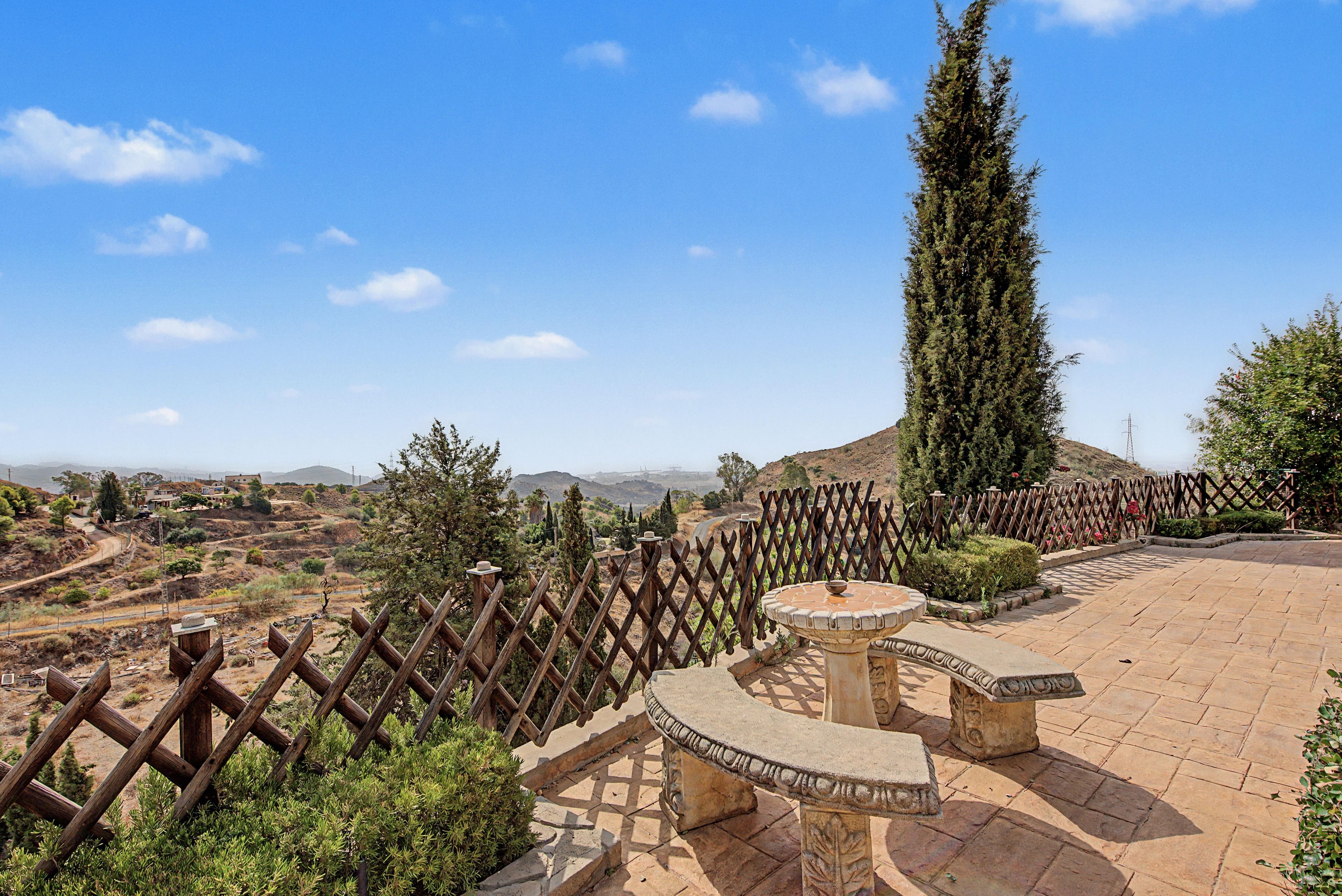 A courtyard with a stone fountain, a wooden fence, and a view of distant hills.