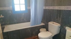 A bathroom with a bathtub, two toilets, and a window with a blue curtain.