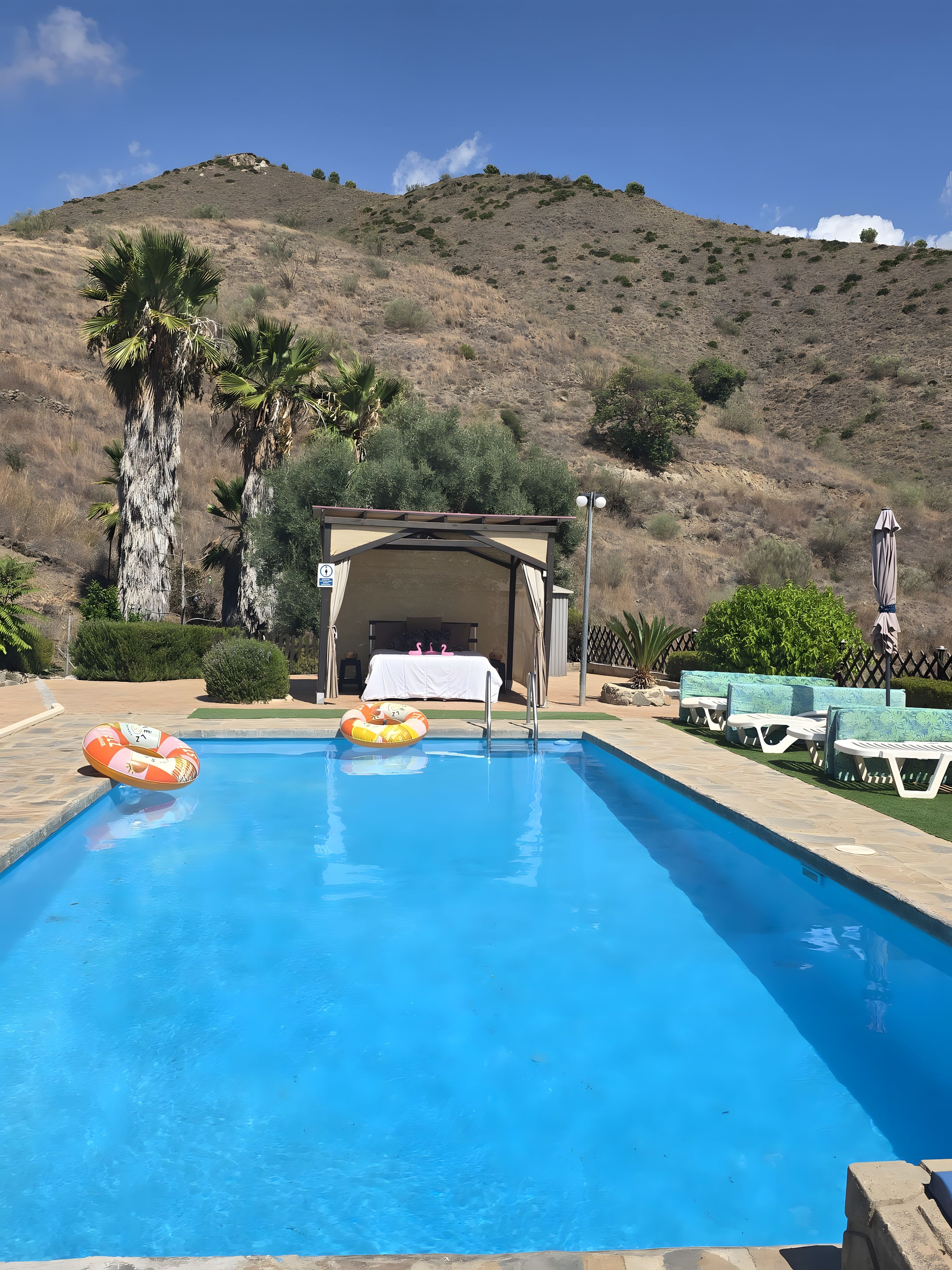 A swimming pool with lounge chairs and a covered area, surrounded by hills.
