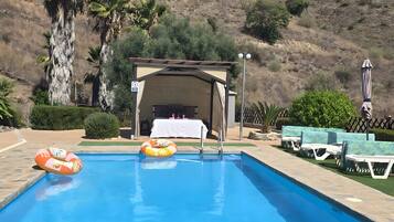 A swimming pool with lounge chairs and a covered area, surrounded by hills.