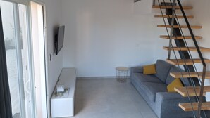 Ein modernes Wohnzimmer mit grauem Sofa, einer weißen Bank, einem kleinen runden Tisch, einem an der Wand befestigten Fernseher, einer Korbtarnlampenschirm und einer Holztreppe.