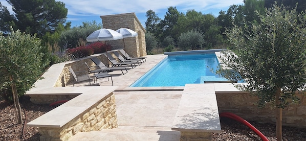Une piscine entourée de murs de pierre et de verdure, avec des chaises longues et des parasols.
