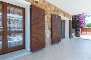 Eine Mauer aus Stein mit hölzernen Fensterläden, einer gefliesten Terrasse und einer blühenden, violetten Pflanze.