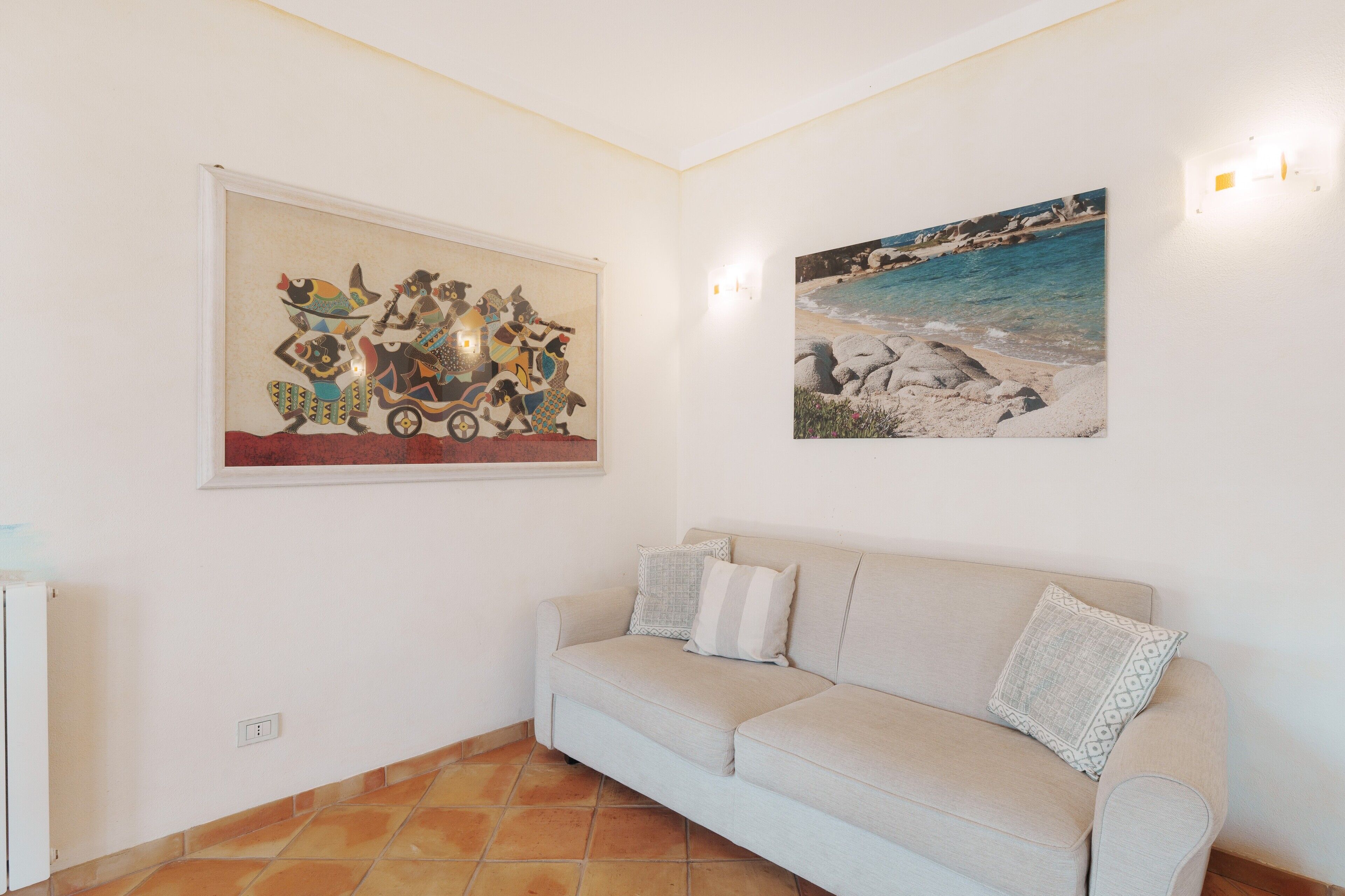 A living room with a beige sofa, two wall-mounted paintings, and a tiled floor.