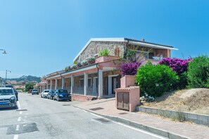 Un edificio con facciata in pietra, un balcone adornato da fiori viola e auto parcheggiate lungo la strada.