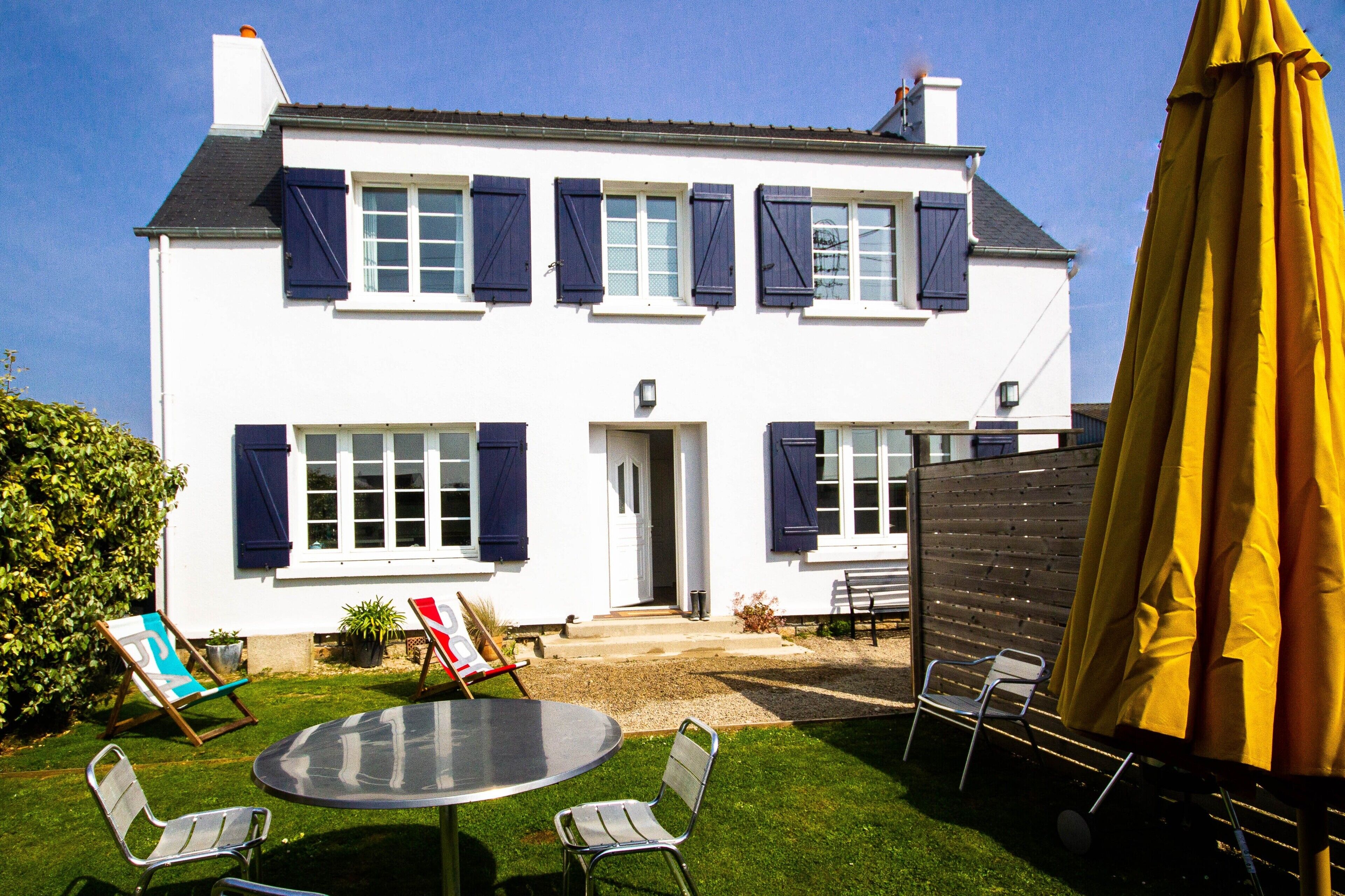 Une maison à deux étages, avec une façade blanche, des volets sombres, une porte d’entrée et un petit jardin devant, agrémenté d’une table et de chaises.