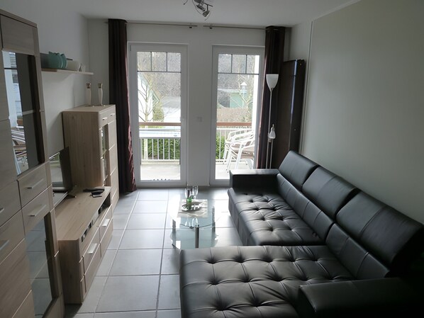 Ein modernes Wohnzimmer mit einer schwarzen Ledersofa, einem Glastisch und einer Schiebetür, die auf einen Balkon führt.
