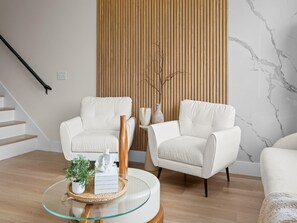 Ein modernes Wohnzimmer mit zwei weißen Sesseln, einem Glastisch und einer Wandpaneele aus Holz.