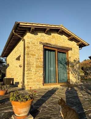 Ein Steinhaus mit einer Holztür und Fensterläden, einem kleinen Sitzbereich im Freien und einer Katze auf dem Boden.