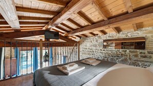 Un plafond en bois, des murs en pierre, un lit avec une couverture et une vue à travers la rambarde d’un balcon.