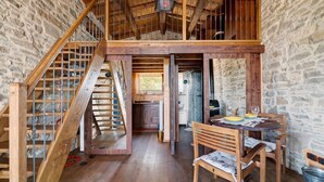Un intérieur rustique avec un escalier en bois, un coin repas comprenant une table et des chaises, et une cuisine visible à travers une porte ouverte.
