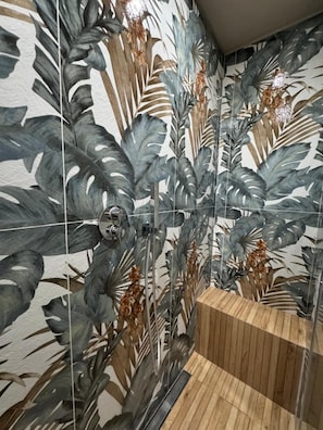 Een badkamer met een douchegedeelte, een houten bankje en een betegelde muur met een bladmotief.