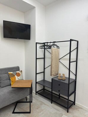 Een kamer met een grijze bank, een houten bijzettafeltje, een zwarte metalen plankkast met een hangende kledingstuk en schoenen, en een flatscreen televisie aan de wand.