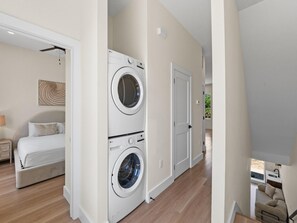 Ein Waschraum mit zwei übereinander gestapelten Waschmaschinen und Trocknern, ein Bett mit Kopfteil sowie ein Flur, der zu einer Tür führt.