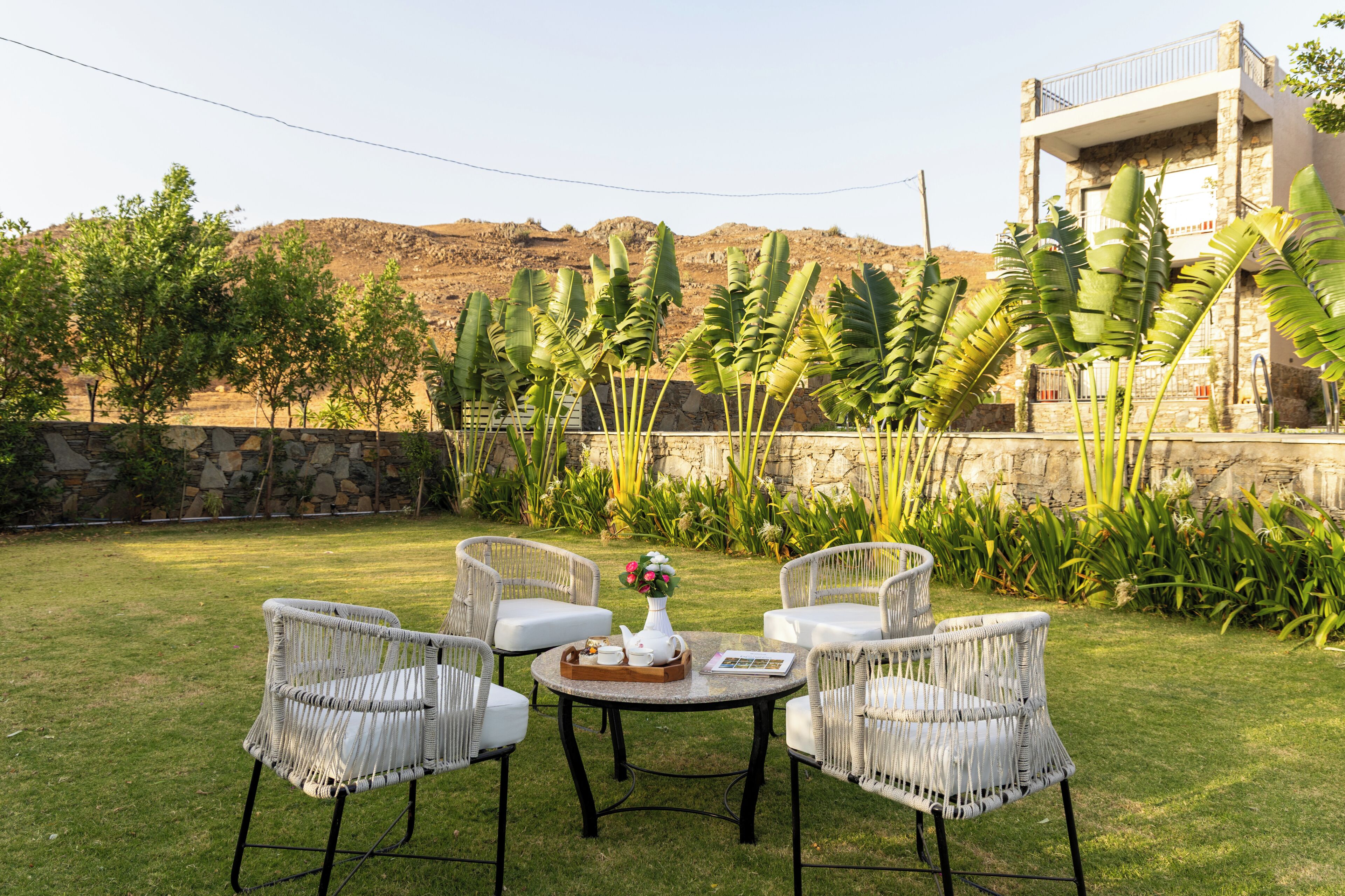 An outdoor seating area with wicker chairs and a table, surrounded by greenery and a stone wall.