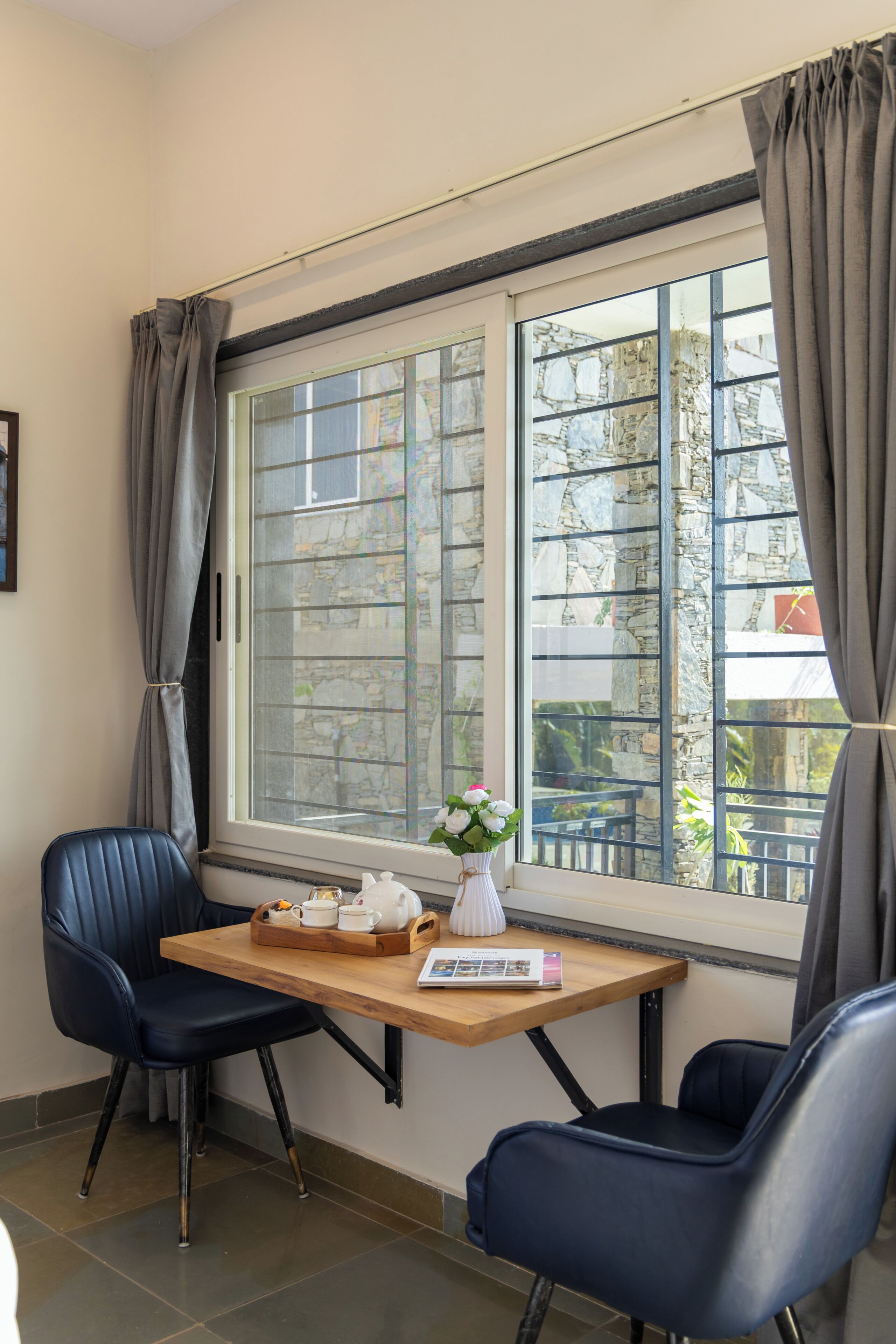 A room with a wooden table, two chairs, a window with curtains, and a vase with flowers.