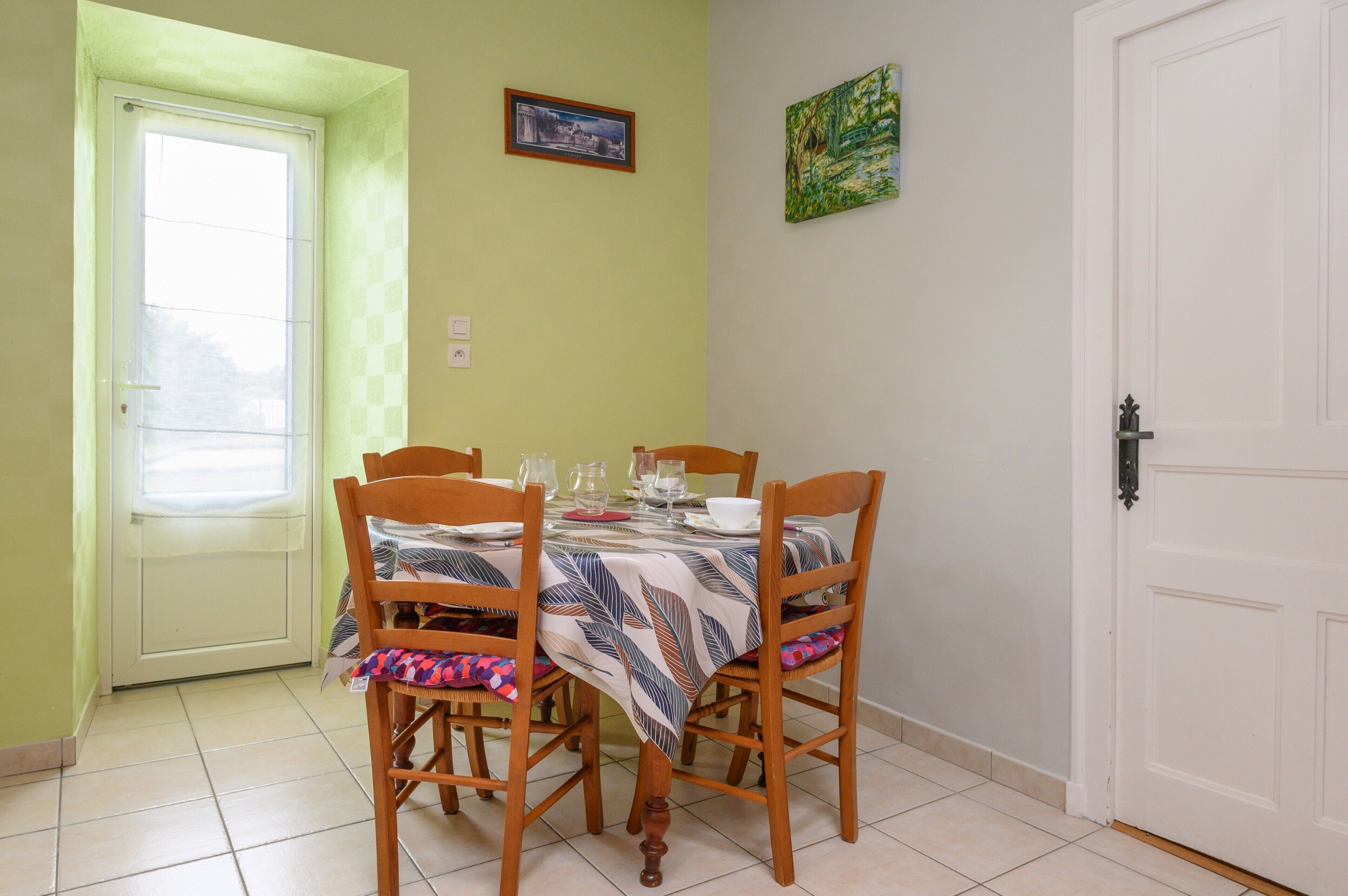 A dining area with a table set for four, wooden chairs, a door, and two framed pictures on the wall.