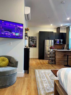 A modern living room with a sofa, a flat-screen TV, a kitchen area with a refrigerator and microwave, and a dining area with a bar stool. - Forest Breeze Studio, Downtown with King Bed (Halifax)