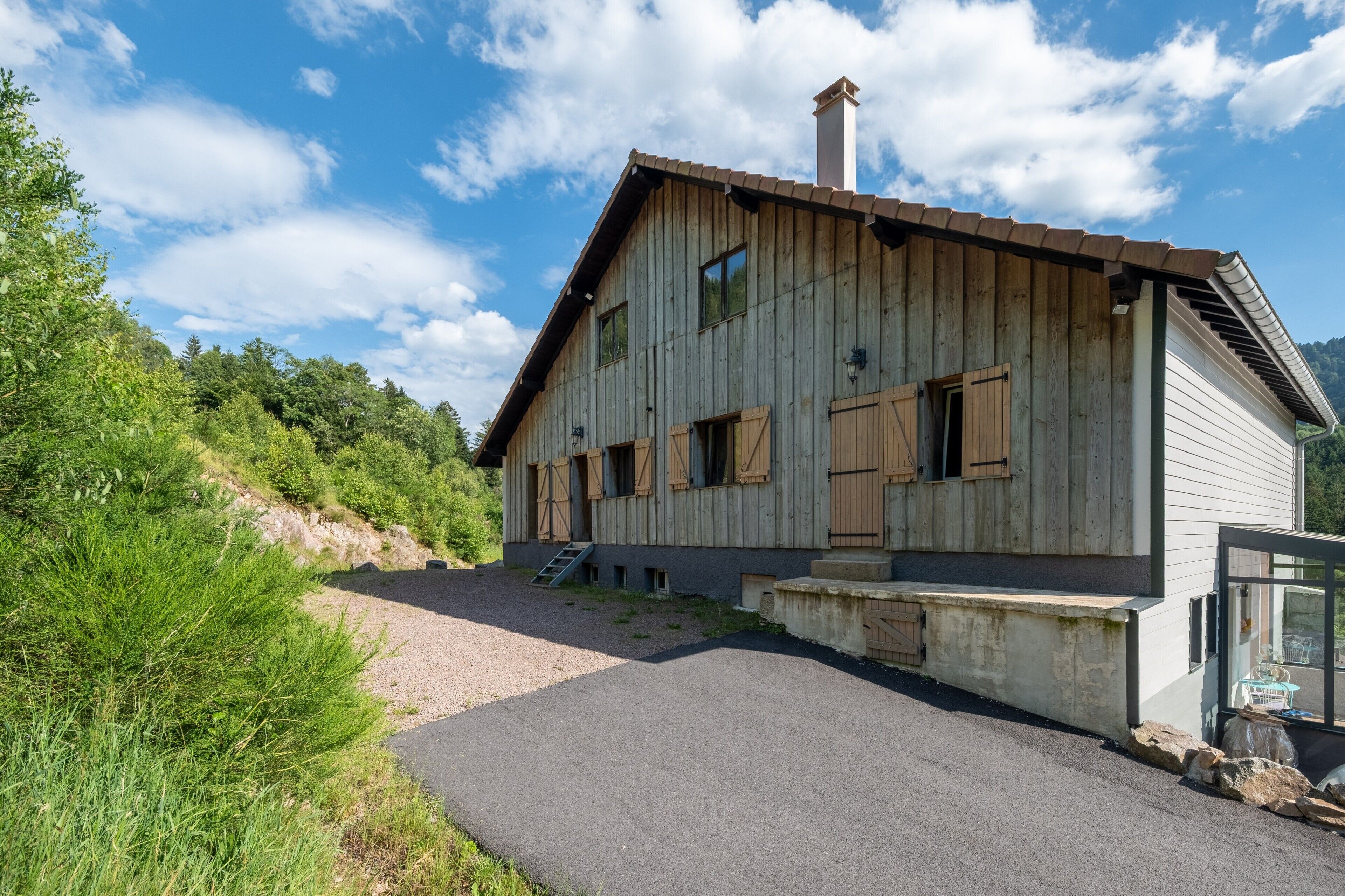 A wooden house with a sloped roof, a chimney, and a gravel driveway leading to a wooden staircase.