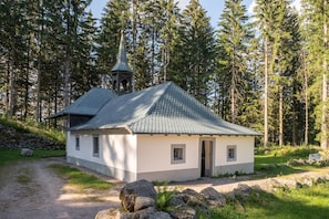 A small, single-story building with a sloped roof, a bell tower, and a stone wall in a forested area.