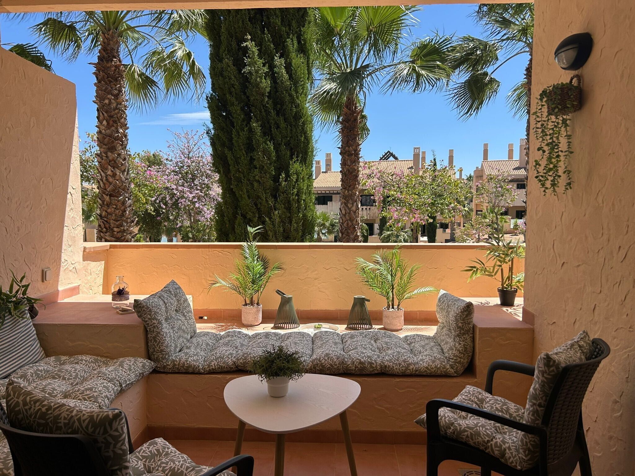 Een zonnig terras met een bank, stoelen en een tafel, omgeven door potplanten en palmbomen.