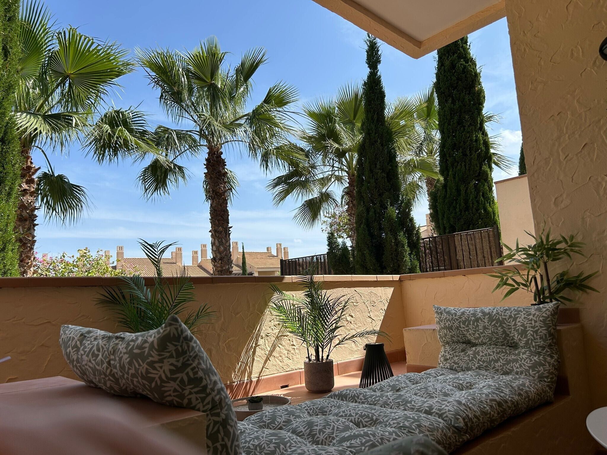 Ein Balkon mit Sitzgelegenheit, Palmen und Blick auf Gebäude.