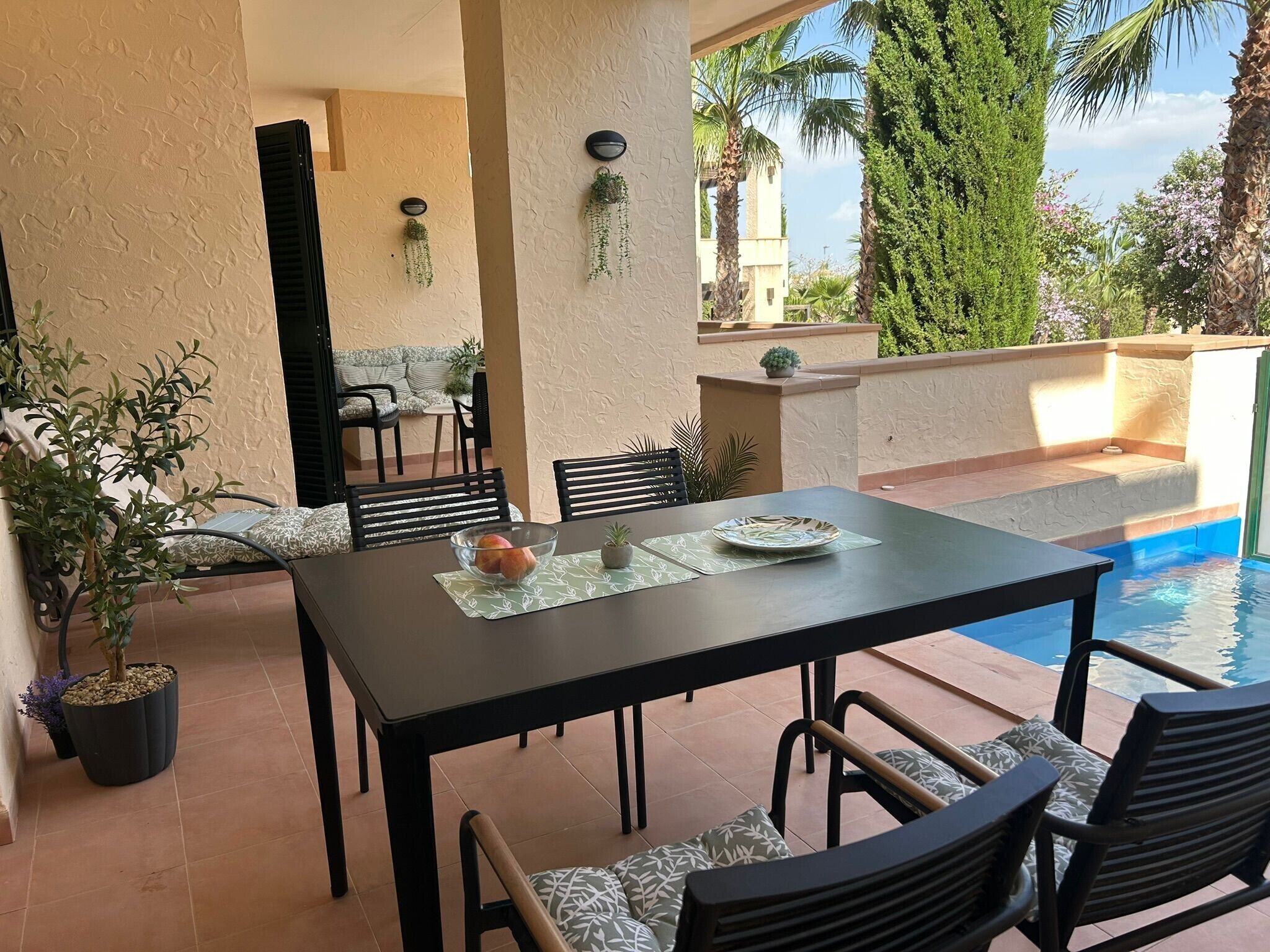 Un área de comedor al aire libre con piscina, rodeada de plantas y una vista despejada del cielo.