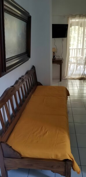 Une chambre avec un lit en bois, un téléviseur fixé au mur, une petite table avec une lampe et une fenêtre ornée de rideaux légers.
