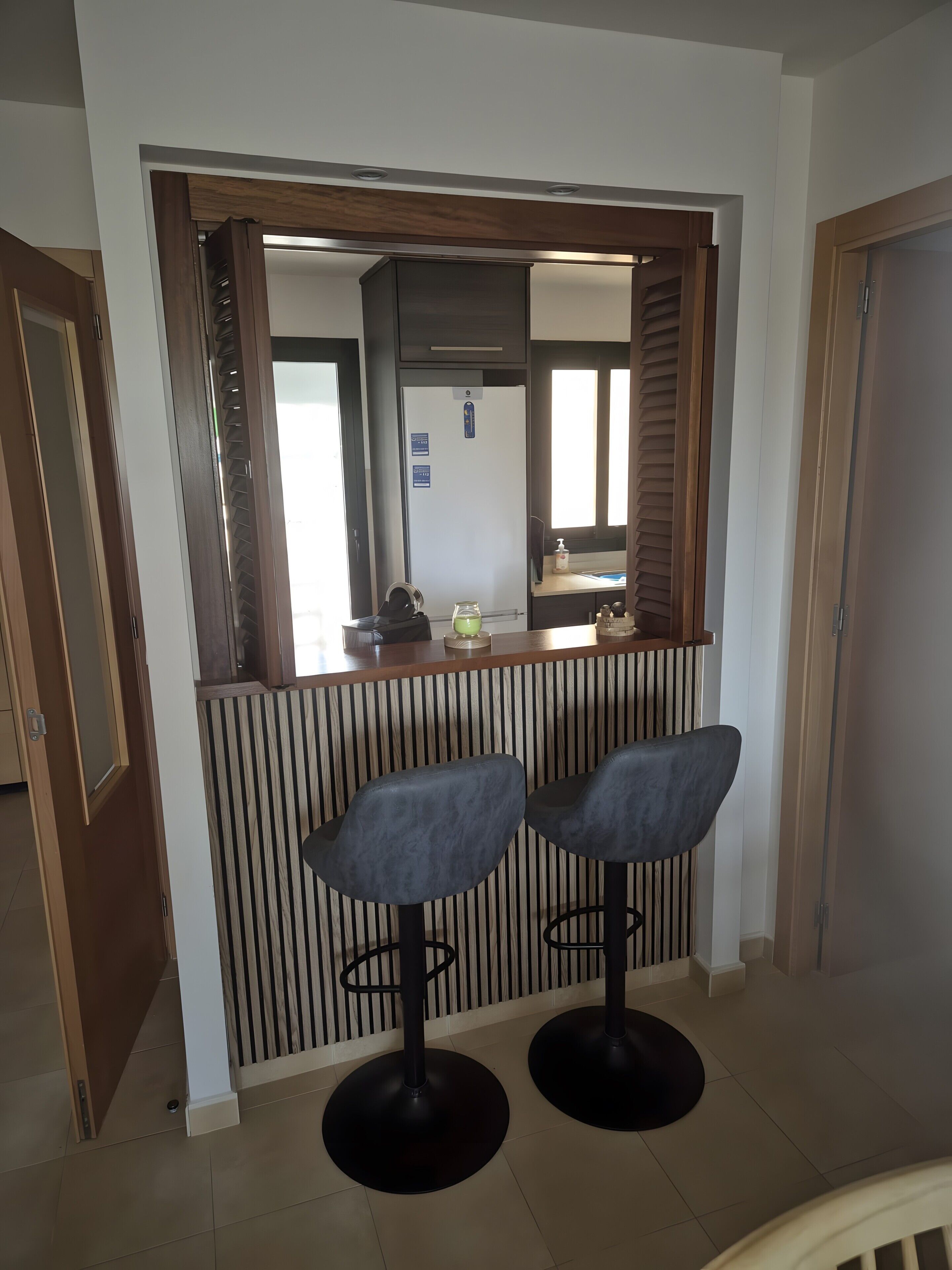 A bar area with two stools, a refrigerator, and a window with blinds.