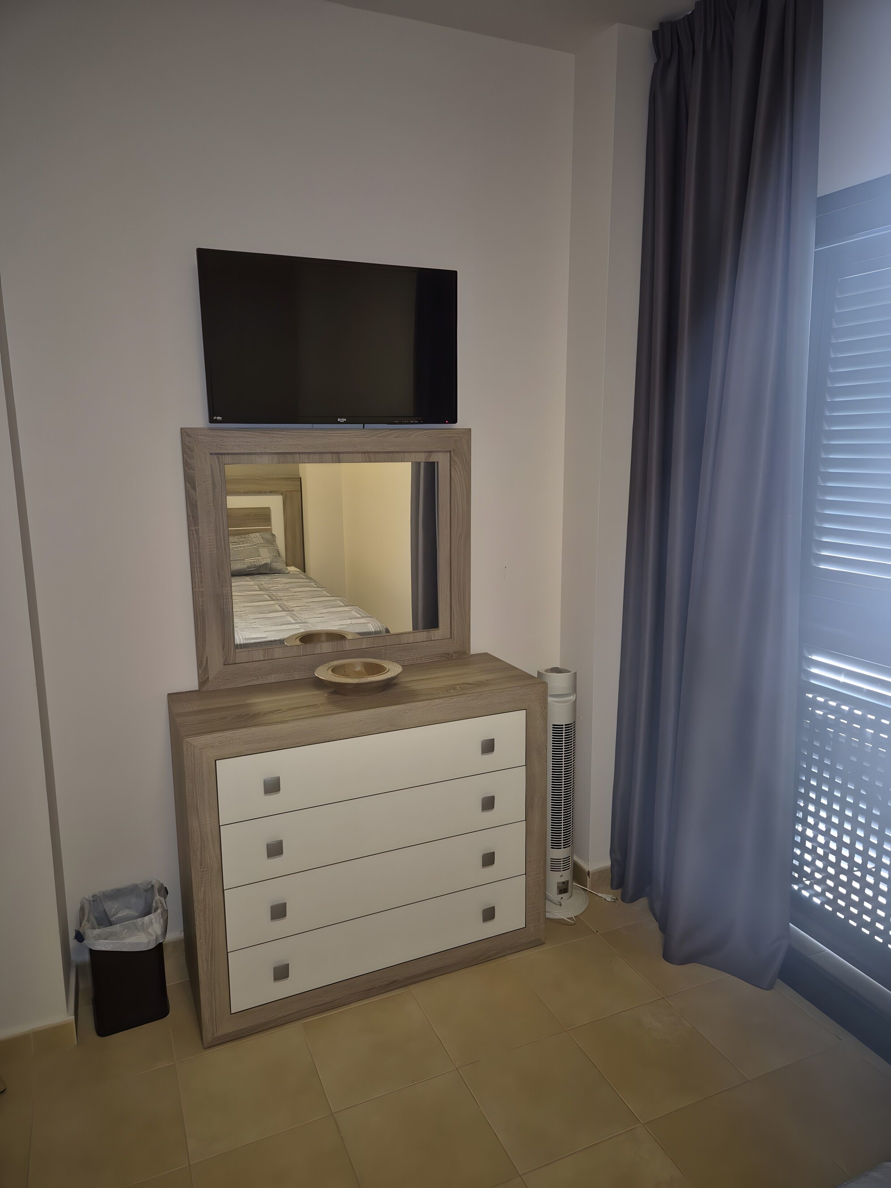 A room with a wooden dresser, a television mounted above it, a mirror, and a window with curtains.