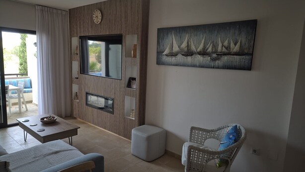 A living room with a sofa, a dining table, a chair, a TV, and a painting of sailboats.