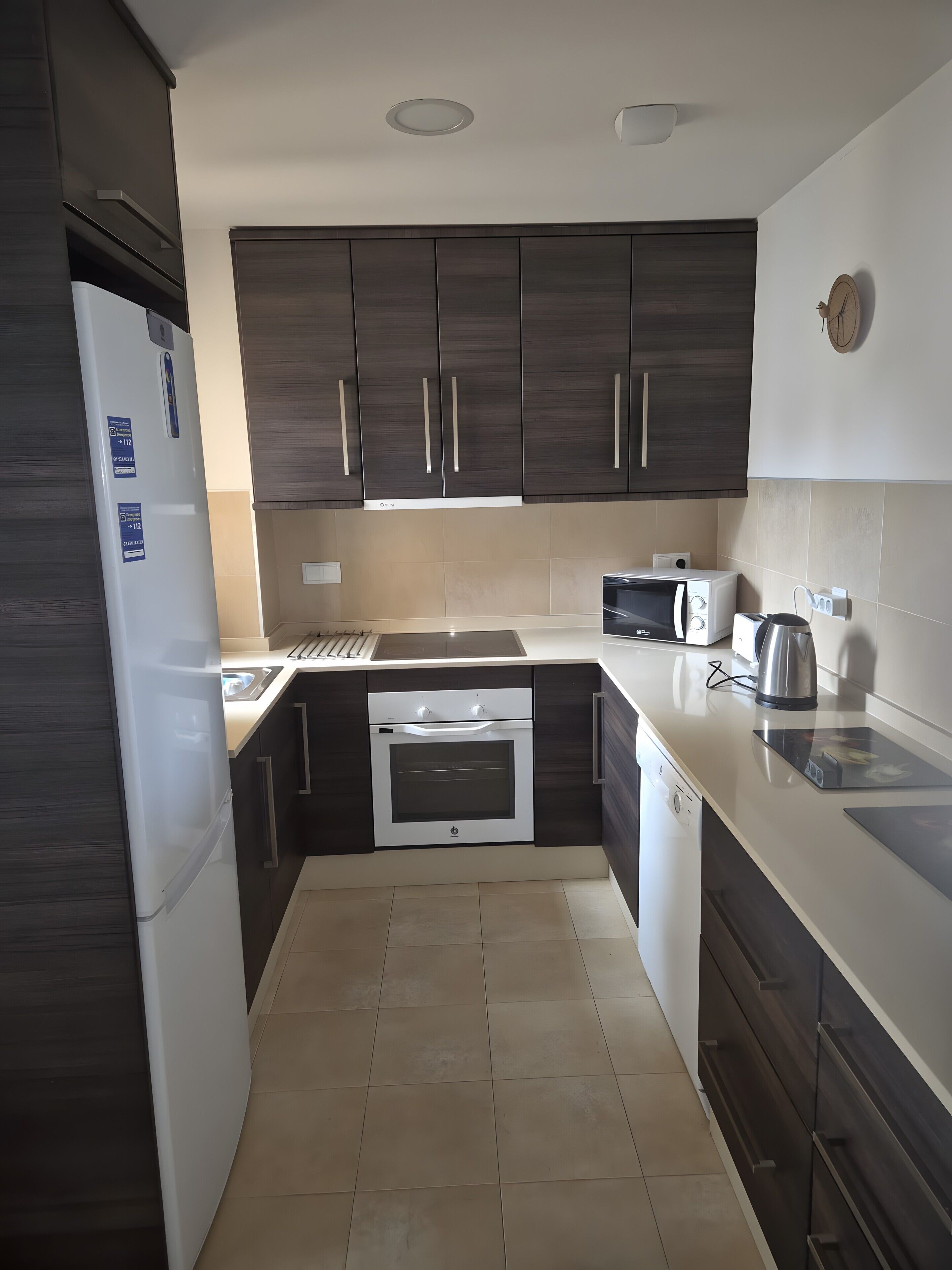 A modern kitchen with dark wood cabinets, a white refrigerator, a built-in oven, a microwave, and a kettle on the countertop.