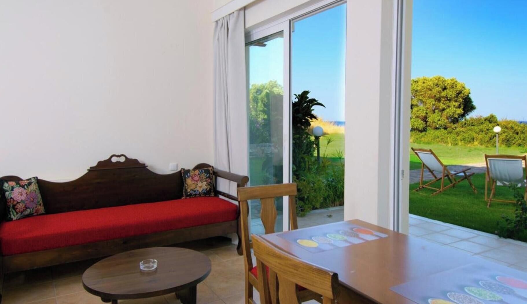 Une pièce avec un canapé rouge, une petite table ronde et une vue sur un espace verdoyant où se trouvent une chaise et une lampe à l’extérieur.