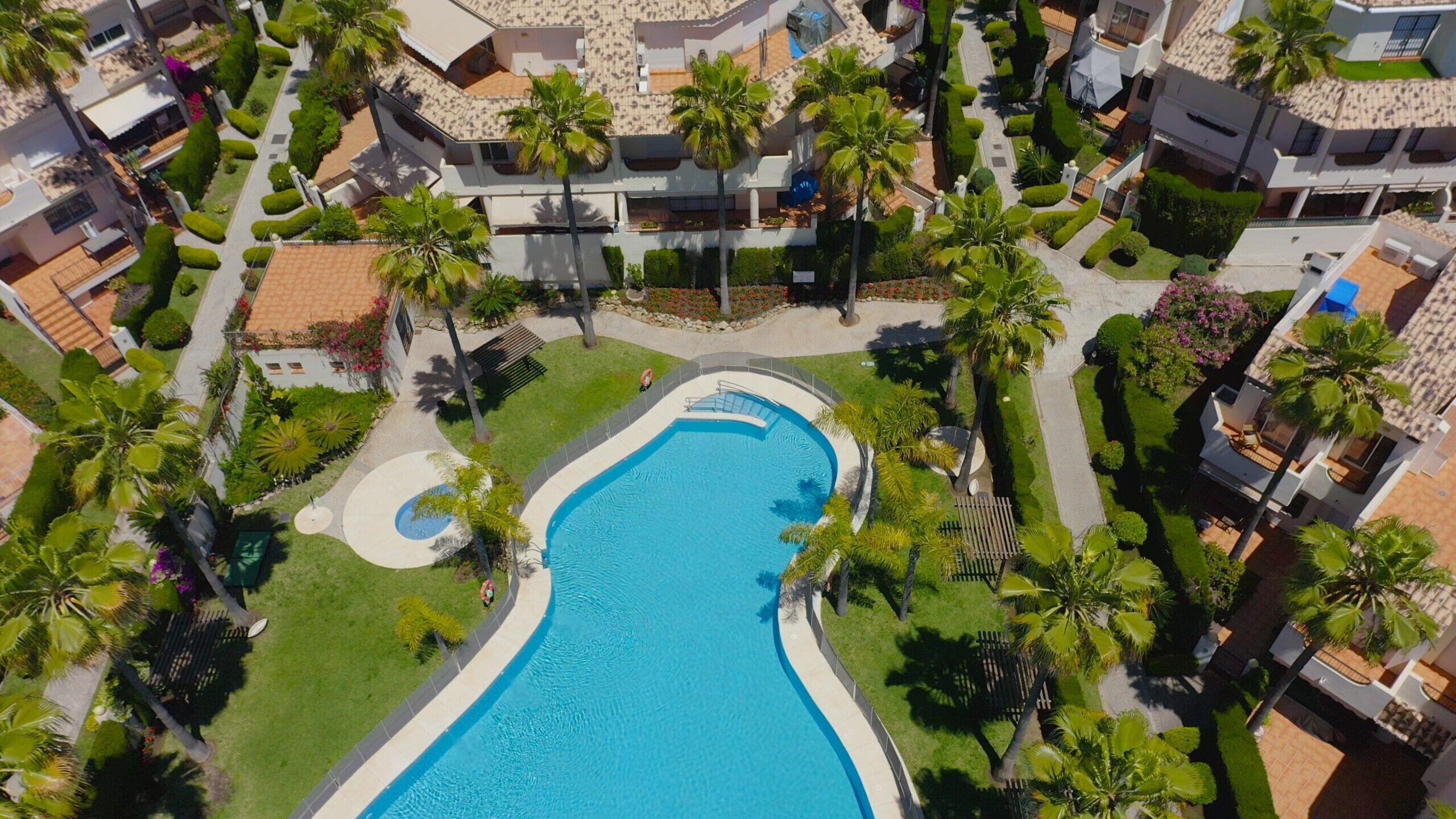 Vue aérienne d’un complexe hôtelier doté d’une grande piscine, entourée de palmiers et de pelouses bien entretenues.