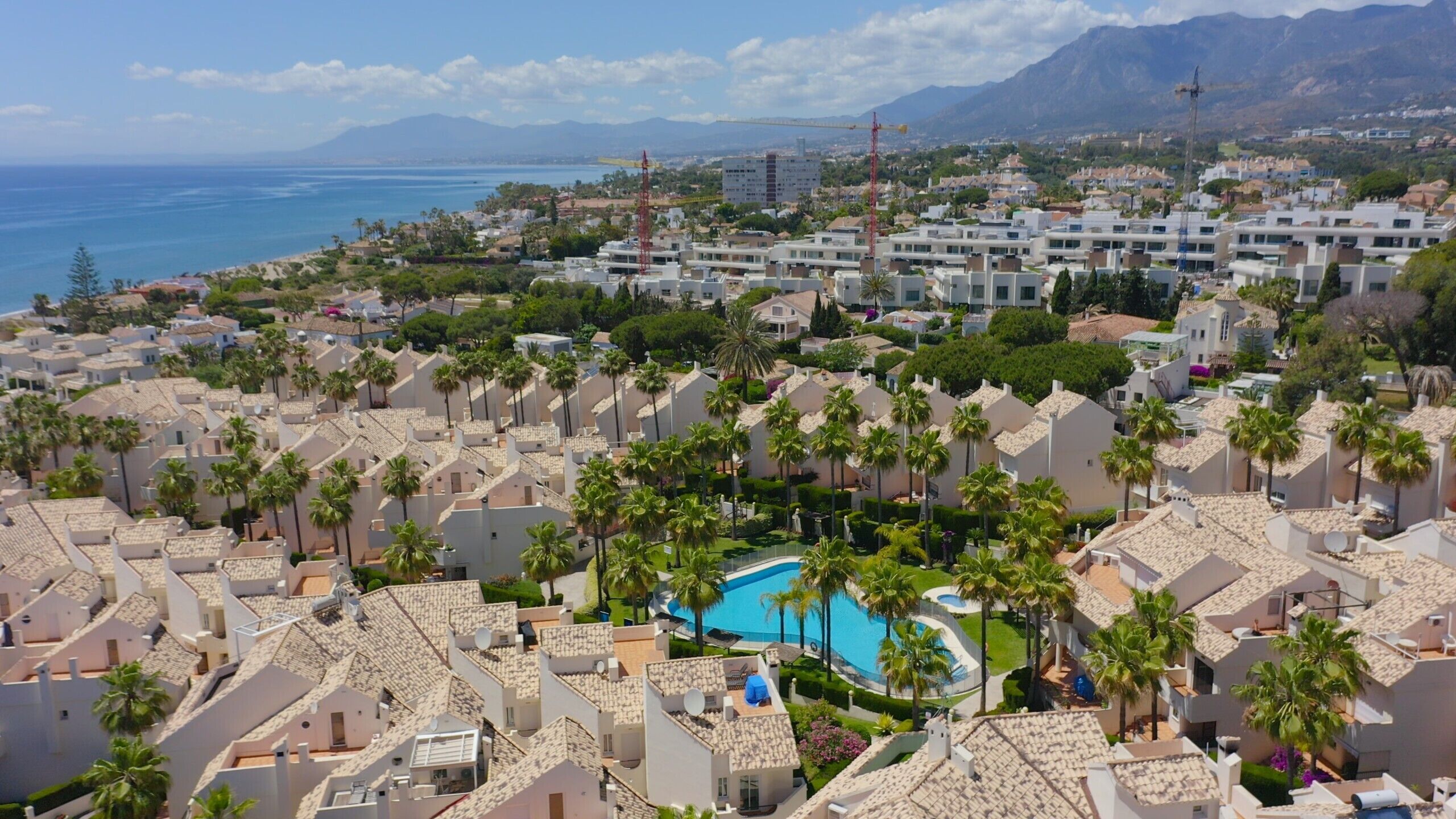 Ein Küstenresort mit mehreren Gebäuden, einem zentral gelegenen Swimmingpool, der von Palmen umgeben ist, und Blick auf das Meer und die Berge im Hintergrund.